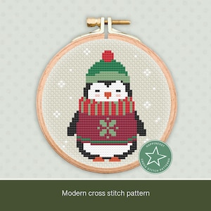 May include: A cross stitch pattern featuring a penguin wearing a red and green sweater and a green hat. The penguin is standing on a white background with snowflakes.