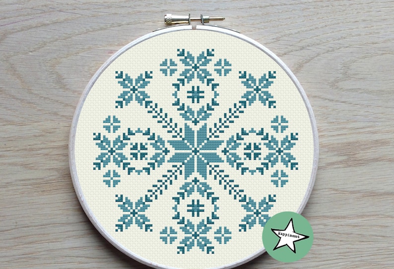 May include: A cross-stitch embroidery hoop featuring a teal and off-white snowflake design. The design is centered on the off-white fabric and framed by a wooden hoop. The snowflake is composed of geometric shapes and floral elements.