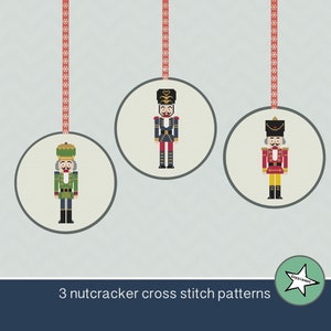 Puede incluir: Tres patrones de punto de cruz de cascanueces, cada uno en un círculo con una cinta roja colgando de la parte superior. Los cascanueces son de diferentes colores: verde, azul y rojo.