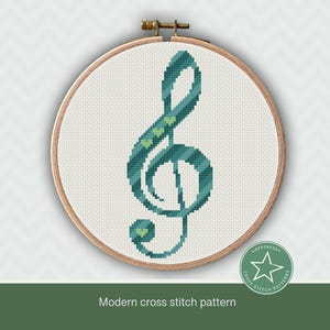 May include: A teal and green cross stitch pattern of a treble clef with small green hearts on the lines. The pattern is framed in a wooden hoop.