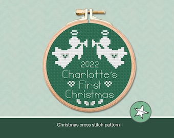 Könnte beinhalten: Ein Kreuzstich-Stickrahmen mit grünem Hintergrund zeigt zwei weiße Engelsfiguren und den Text "2022 Charlotte's First Christmas". Der Holzrahmen ist mit einem Messingverschluss befestigt. Das Design ist ein Weihnachtskreuzstichmuster.