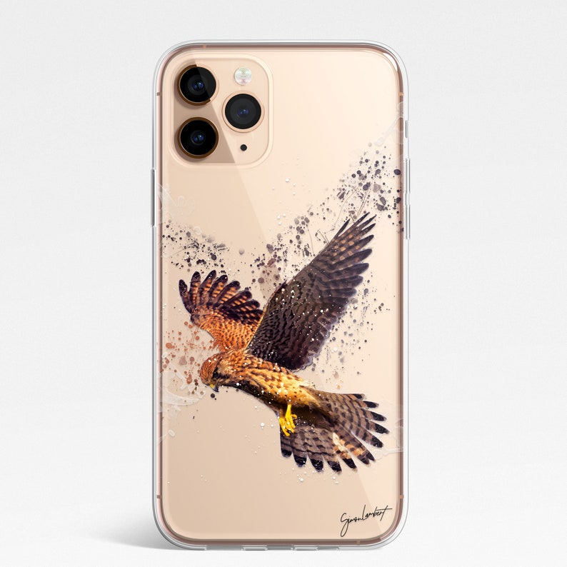 May include: Clear phone case featuring a watercolor illustration of a hawk in flight. The artwork showcases the bird with brown and tan feathers, wings spread, and dark ink splatter effects. The case has a glossy finish and a signature in the bottom right corner.