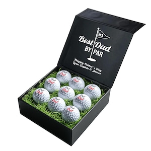 Op de afbeelding: Zwarte geschenkdoos met open klapdeksel, gevuld met witte golfballen met de tekst "Best Dad". De binnenkant van het deksel toont de tekst "Best Dad By Par" en "Happy Father's Day Love Sophie & James". Een cadeau voor Vaderdag.