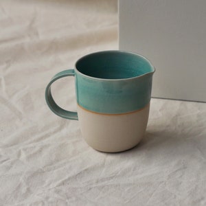 May include: A light blue and white ceramic pitcher with a rounded shape and a handle. The pitcher has a light blue glaze on the top half and a white glaze on the bottom half.