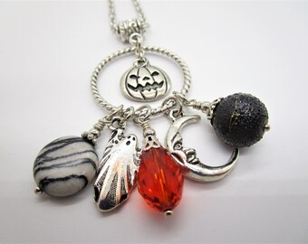 May include: A silver necklace with a Halloween theme. It features a jack-o'-lantern charm, a crescent moon charm, a ghost charm, a red glass bead, a black lava rock bead, and a white and black striped stone bead.