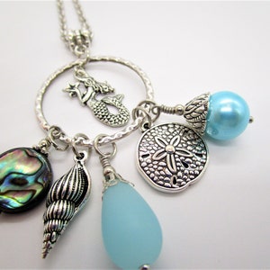 May include: Silver necklace with a mermaid charm, a seashell charm, a sand dollar charm, a blue glass bead, and a blue abalone shell charm.
