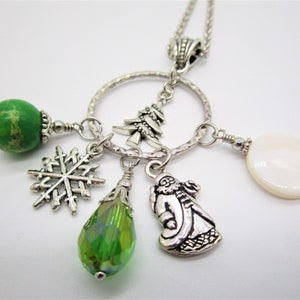 Puede incluir: Un collar de plata con un colgante circular. El colgante tiene un dije de copo de nieve, un dije de cristal verde, un dije de Santa Claus, una cuenta de piedra verde, una cuenta de concha blanca y un dije de árbol de Navidad plateado.