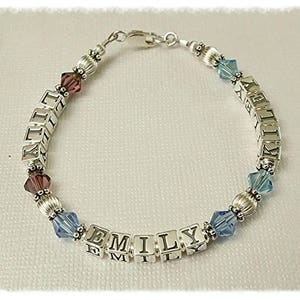 May include: A silver bracelet with the names "EMILY" and "KELLEY" spelled out in square letter beads. The bracelet features purple and blue crystal beads and silver accents. The bracelet is designed with a clasp closure.