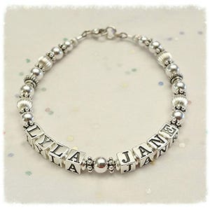 May include: Silver-toned bracelet featuring the names "LYLA" and "JANE" spelled out on square letter beads. The bracelet includes various silver beads and decorative elements, creating a personalized and elegant accessory.