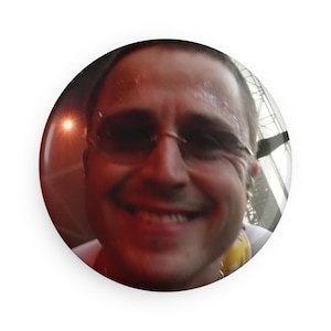 May include: A circular pinback button featuring a photographic portrait of a smiling person wearing dark-tinted rimless glasses. The image has a warm, slightly blurry quality with a bright light source visible in the background on the left side.