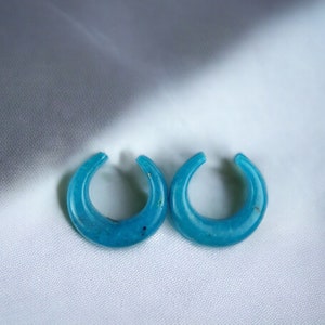 May include: A pair of turquoise crescent-shaped earrings. The earrings are a bright blue-green hue, with a smooth, polished finish. The earrings are presented on a white, textured surface.