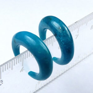 May include: Two turquoise-coloured, curved earrings. The earrings are a vibrant blue-green hue, with a smooth, polished surface. The earrings are placed on a ruler, showing their size. The earrings are a bold and stylish accessory.