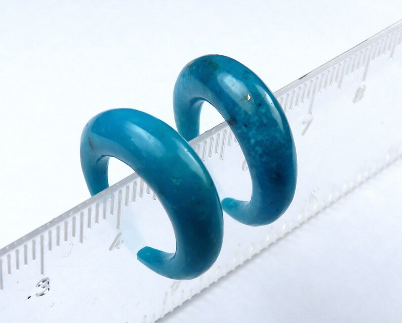 May include: Two turquoise-coloured, curved earrings. The earrings are a vibrant blue-green hue, with a smooth, polished surface. The earrings are placed on a ruler, showing their size. The earrings are a bold and stylish accessory.