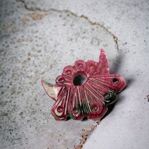 Op de afbeelding: Een gebeeldhouwde, roze edelsteen hanger met een bloemmotief. De hanger heeft een centrale cirkelvormige opening, bloembladachtige vormen en ingewikkelde gesneden details. De edelsteen heeft een mix van roze, witte en groene tinten.