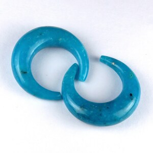 May include: Two turquoise crescent-shaped earrings. The earrings are a vibrant blue-green hue, with a smooth, polished surface. The curved design is connected, creating a unique and eye-catching look. The earrings are set against a white background.