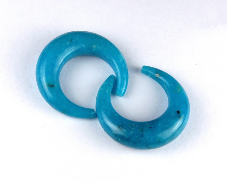 May include: Two turquoise crescent-shaped earrings. The earrings are a vibrant blue-green hue, with a smooth, polished surface. The curved design is connected, creating a unique and eye-catching look. The earrings are set against a white background.