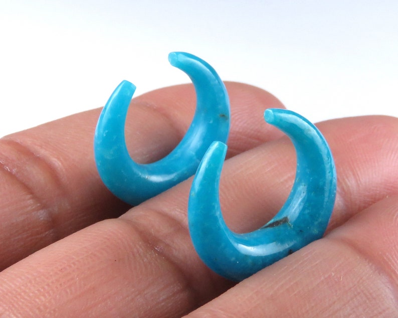 May include: A pair of turquoise crescent-shaped earrings. The earrings are a vibrant blue-green hue, with a smooth, polished surface. The curved design is reminiscent of a horn or a stylized moon. The earrings are shown against a neutral background.