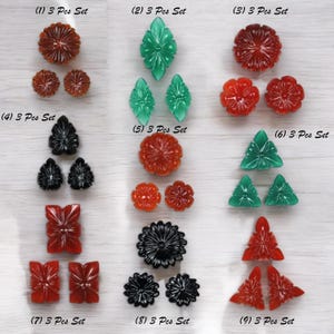 Könnte beinhalten: Eine Sammlung von neun Sets geschnitzter Edelsteinstücke, jeweils als "3 Pcs Set" bezeichnet. Die Sets zeigen verschiedene Blumen- und geometrische Designs in Farben wie Rot, Grün und Schwarz. Die Stücke werden auf einer hellen Holzmaserungsoberfläche präsentiert.