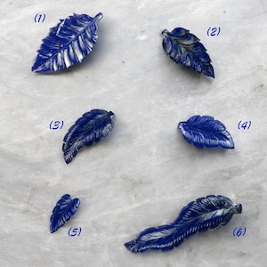 Könnte beinhalten: Sechs geschnitzte Anhänger in Form von Lapislazuli-Blättern in verschiedenen Größen. Die Blätter sind tiefblau mit weißen Adern und auf einer hellgrauen Oberfläche angeordnet. Die Blätter sind aufwendig detailliert und zeigen die natürliche Schönheit des Steins.
