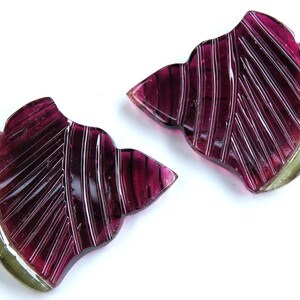 May include: Two carved gemstone slices in a leaf shape. The gemstones are a deep magenta color with etched lines and a pale green edge. The gemstones are set against a white background.