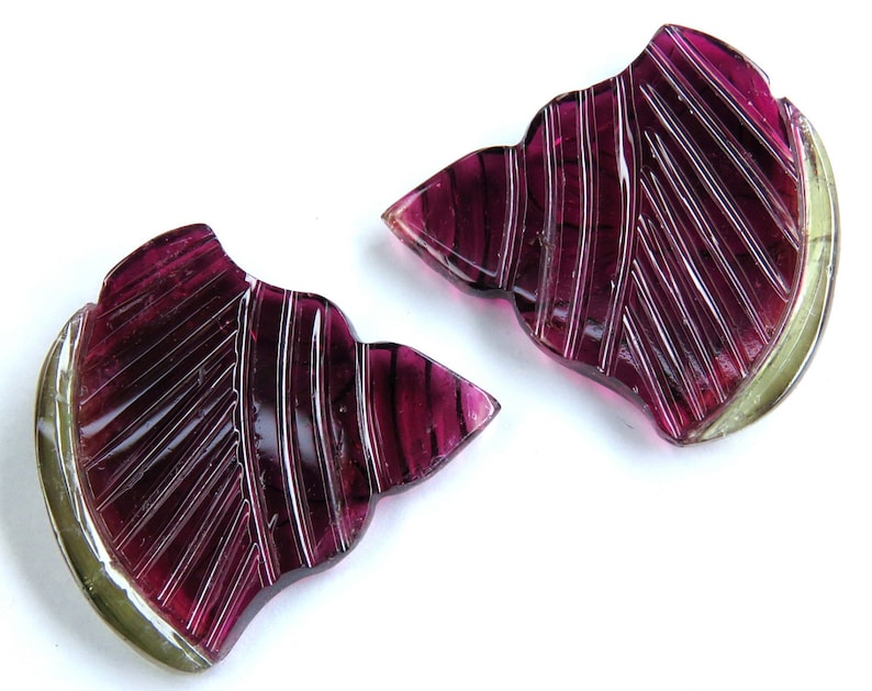 May include: Two carved gemstone slices in a leaf shape. The gemstones are a deep magenta color with etched lines and a pale green edge. The gemstones are set against a white background.