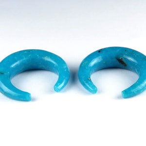 May include: A pair of turquoise crescent-shaped ear gauges. The gauges are smooth and polished, with a curved design. The open ends are rounded. The background is white. Each gauge is approximately 2.5 cm in diameter.