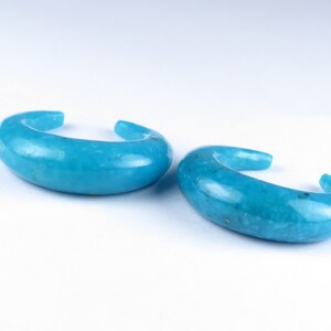 May include: Two turquoise ear cuffs. Each cuff has a curved, open design. The smooth, polished surface of the jewellery reflects light, highlighting the vibrant blue-green hue. The cuffs are displayed against a white background.