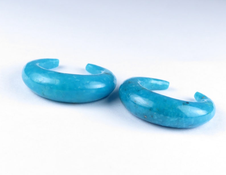 May include: Two turquoise ear cuffs. Each cuff has a curved, open design. The smooth, polished surface of the jewellery reflects light, highlighting the vibrant blue-green hue. The cuffs are displayed against a white background.