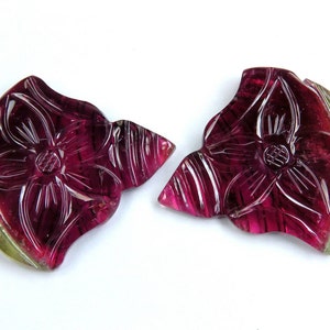 May include: Two carved gemstone pendants in the shape of a flower. The pendants are a deep magenta color with carved details and a green edge. The pendants are on a white background.