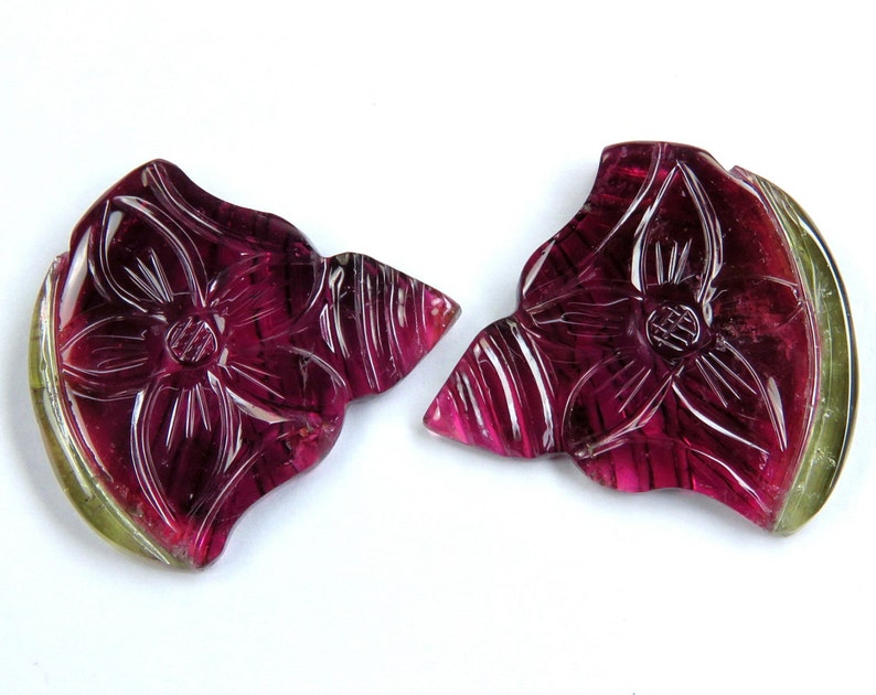 May include: Two carved gemstone pendants in the shape of a flower. The pendants are a deep magenta color with carved details and a green edge. The pendants are on a white background.