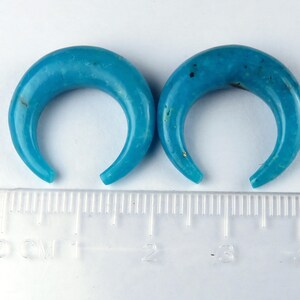 May include: Two turquoise crescent-shaped ear gauges. The gauges are a vibrant blue with natural variations in colour. A ruler with centimetre markings is in the foreground.