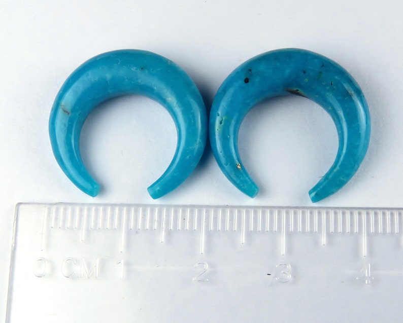May include: Two turquoise crescent-shaped ear gauges. The gauges are a vibrant blue with natural variations in colour. A ruler with centimetre markings is in the foreground.
