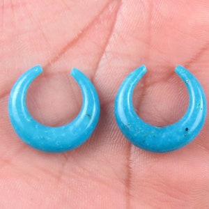 May include: Two turquoise crescent-shaped earrings. Each earring has a smooth, polished surface and a small opening at the top. The earrings are displayed against a neutral background.