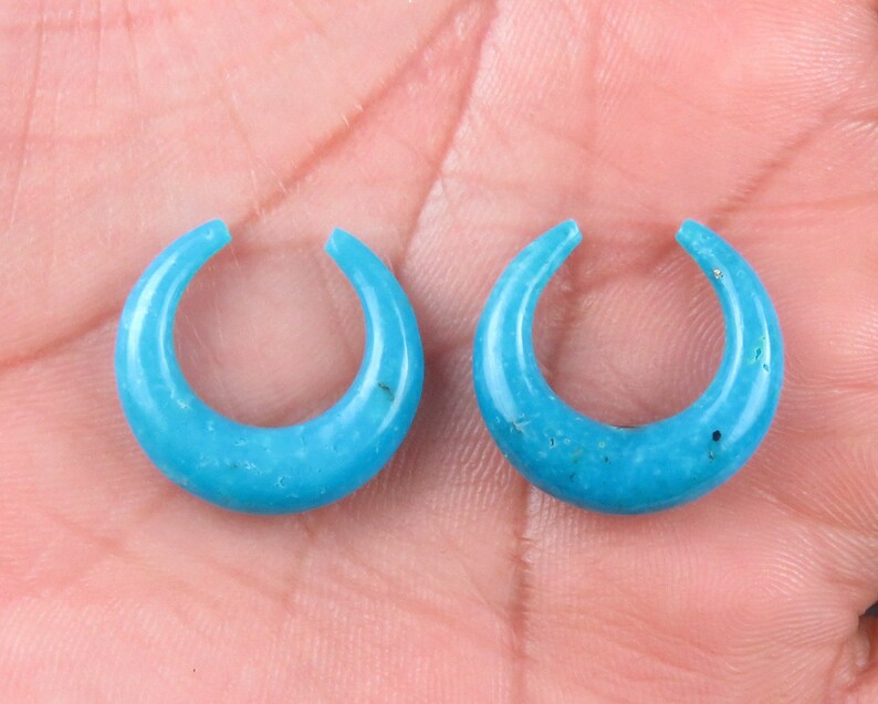 May include: Two turquoise crescent-shaped earrings. Each earring has a smooth, polished surface and a small opening at the top. The earrings are displayed against a neutral background.