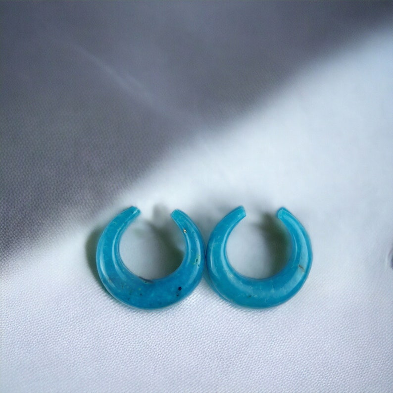 May include: A pair of turquoise crescent-shaped earrings. The earrings are a bright blue-green hue, with a smooth, polished finish. The earrings are presented on a white, textured surface.