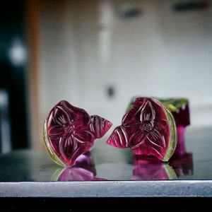 May include: Two carved gemstone flower pendants. The pendants are a deep magenta color with a yellow-green border. The flower design is etched into the stone. The pendants are on a reflective surface.