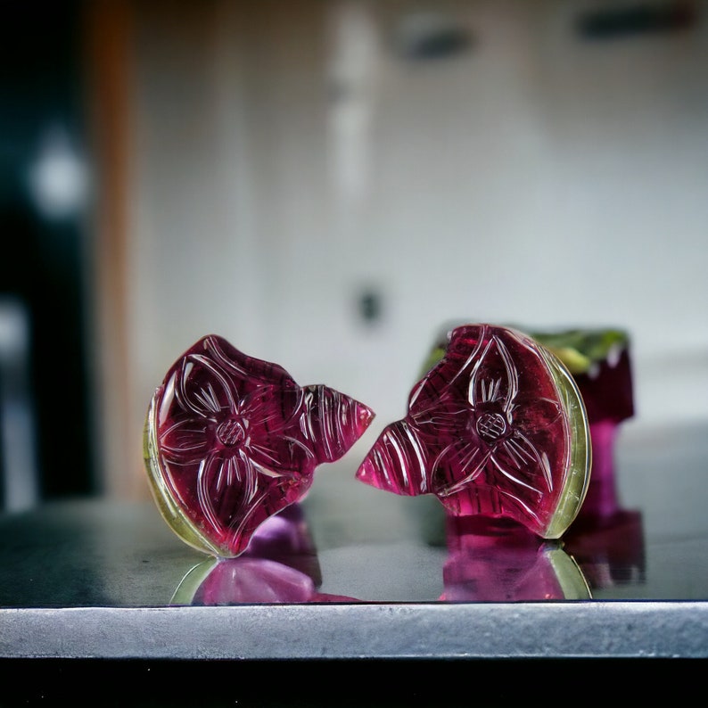 May include: Two carved gemstone flower pendants. The pendants are a deep magenta color with a yellow-green border. The flower design is etched into the stone. The pendants are on a reflective surface.