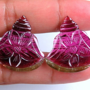 May include: Two carved gemstones in a floral design. The gemstones are a deep magenta color with intricate carved details. The base of the gemstones is a light brown color. The gemstones are held in a person's hand.