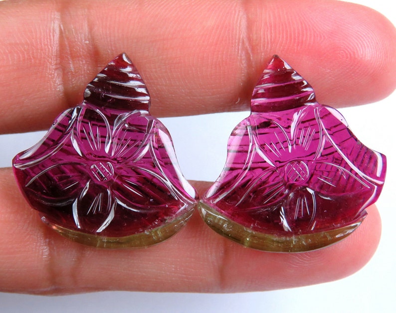 May include: Two carved gemstones in a floral design. The gemstones are a deep magenta color with intricate carved details. The base of the gemstones is a light brown color. The gemstones are held in a person's hand.