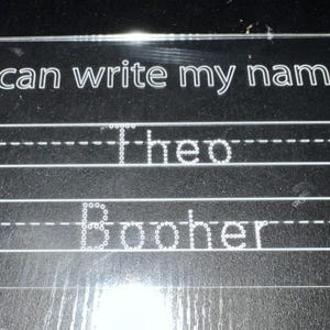 May include: A clear acrylic writing board with the text "I can write my name" at the top. The names "Theo" and "Booher" are written in dotted lines, designed for tracing. The board has horizontal lines for practicing handwriting.