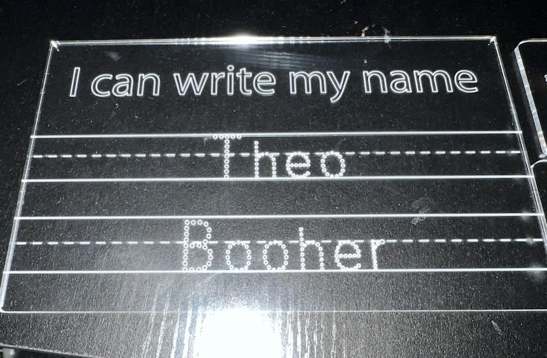 May include: A clear acrylic writing board with the text "I can write my name" at the top. The names "Theo" and "Booher" are written in dotted lines, designed for tracing. The board has horizontal lines for practicing handwriting.