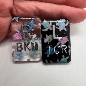 May include: Two rectangular resin keycaps. One is clear with colorful sea turtles and the letters "R" and "BKM" in silver. The other is black with sea turtles and the letters "L" and "TCR" in silver. The keycaps are likely for a keyboard.