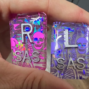 May include: Two rectangular keycaps with a skeleton theme. One is pink with a silver "R" and the text "SAS", the other is purple with a silver "L" and "SAS". Both keycaps feature a skull and holographic accents.