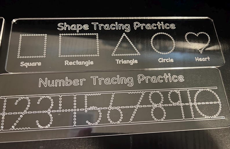 May include: Two clear acrylic tracing boards. The top board says "Shape Tracing Practice" and features dotted outlines of a square, rectangle, triangle, circle, and heart. The bottom board says "Number Tracing Practice" and has dotted numbers 1-10.