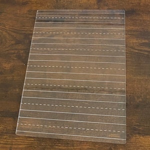 May include: A rectangular writing practice board with a dark brown wood grain pattern. White dashed lines are evenly spaced across the surface, designed for practicing handwriting. The board is made of a clear, durable material.