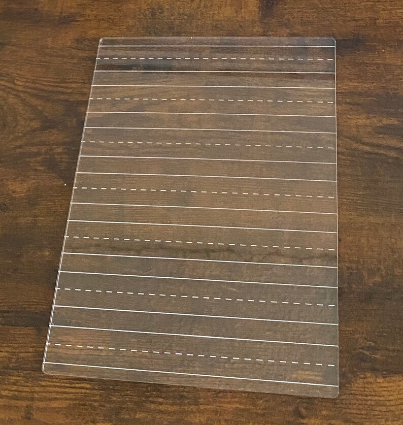 May include: A rectangular writing practice board with a dark brown wood grain pattern. White dashed lines are evenly spaced across the surface, designed for practicing handwriting. The board is made of a clear, durable material.