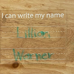 May include: Clear acrylic name tracing board with the text "I can write my name" in white. The names "Lillian" and "Warner" are written in green on the board, following the lines. The board is on a wooden surface.