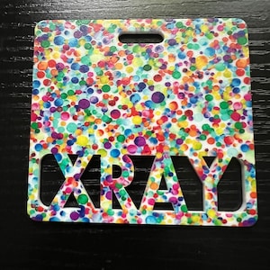 May include: A white plastic badge with a colorful confetti pattern and the word "XRAY" cut out in the center.