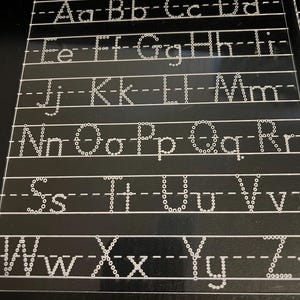 May include: A black acrylic tracing board with the text "I can write my letters" at the top. The board features dotted letters of the alphabet, both uppercase and lowercase, for tracing practice. The letters are in a white, dotted font.