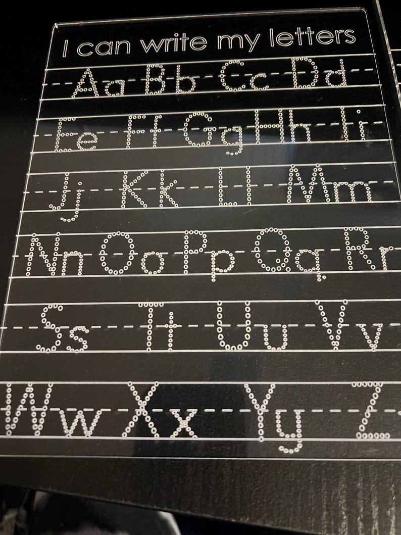May include: A black acrylic tracing board with the text "I can write my letters" at the top. The board features dotted letters of the alphabet, both uppercase and lowercase, for tracing practice. The letters are in a white, dotted font.